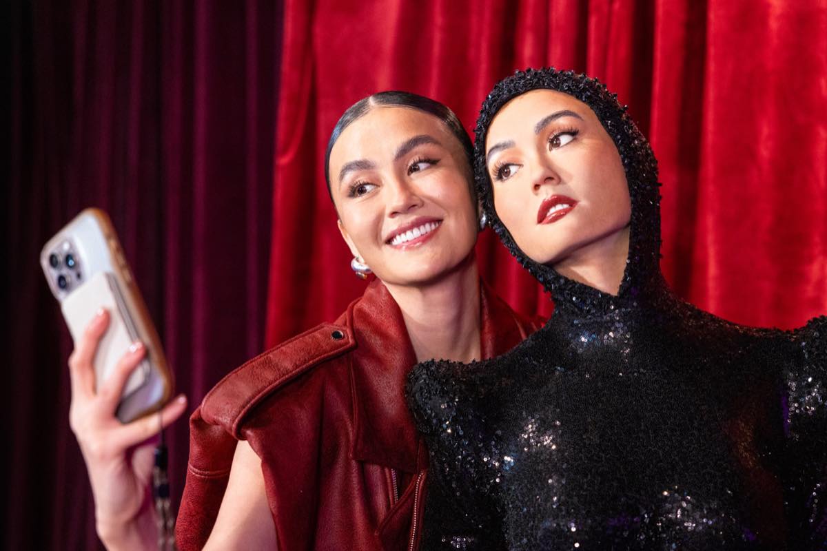 Agnez Mo takes a selfie on a phone beside her seated wax figure against a red backdrop