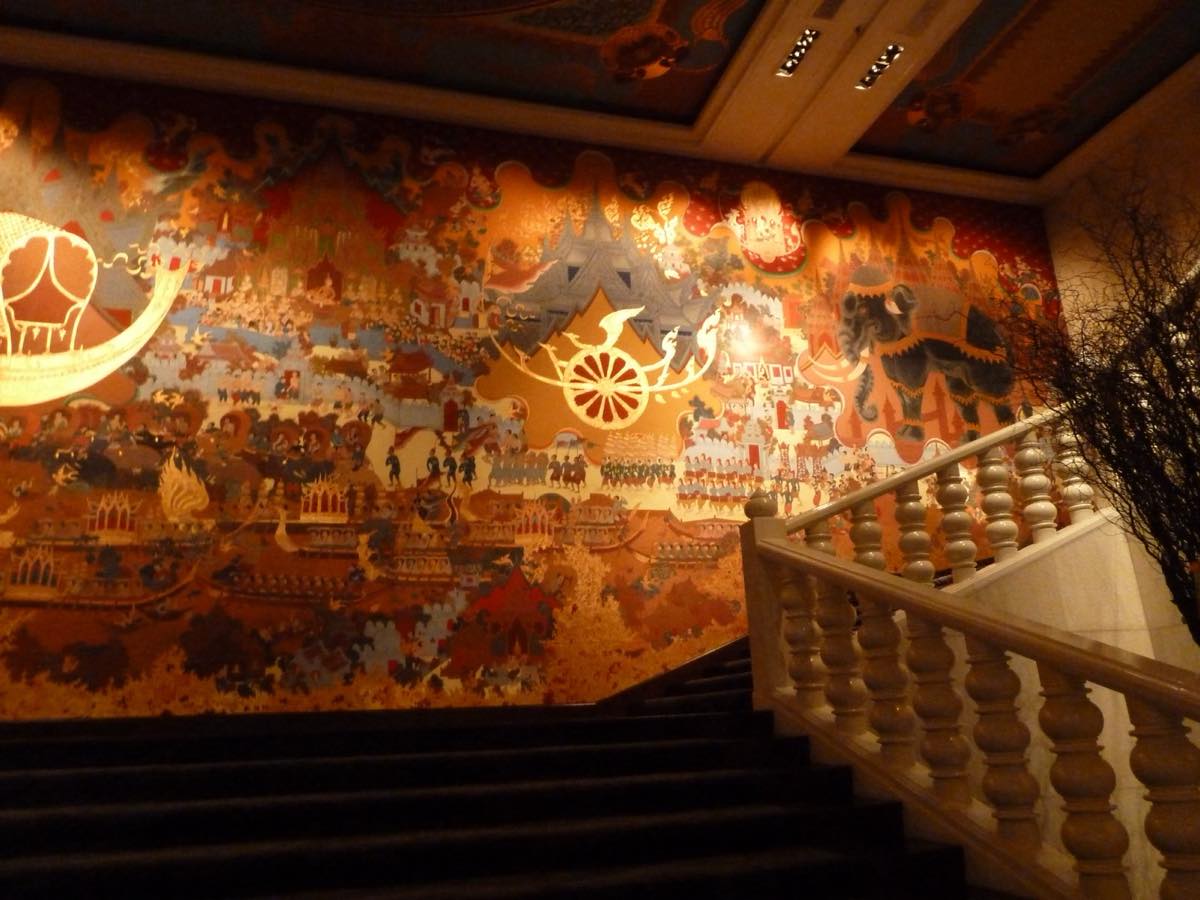The grand lobby staircase at Anantara Siam Bangkok beneath a large hand-painted Thai mural and painted ceiling panels