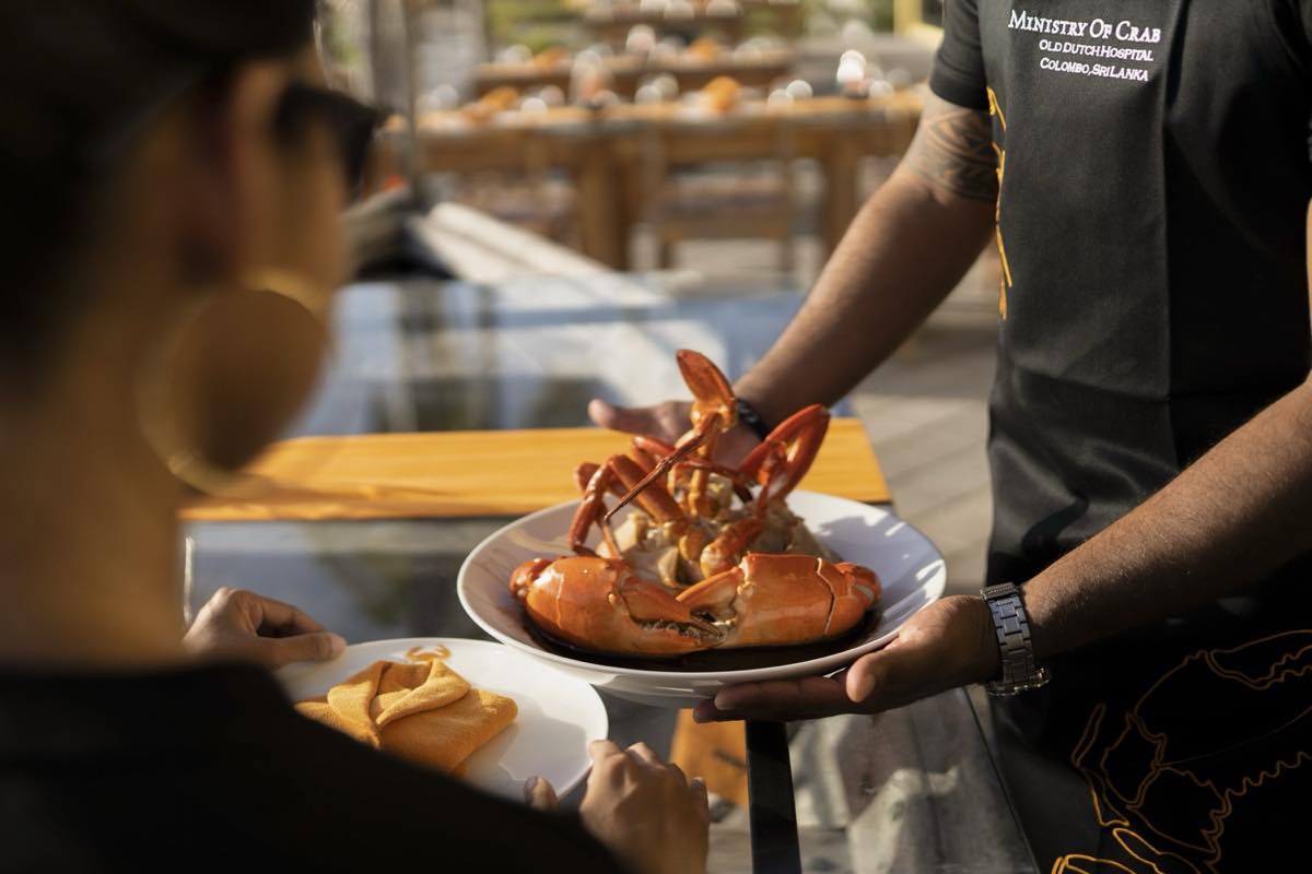 Server in a Ministry of Crab uniform presenting a whole Sri Lankan mud crab on a plate to a seated guest