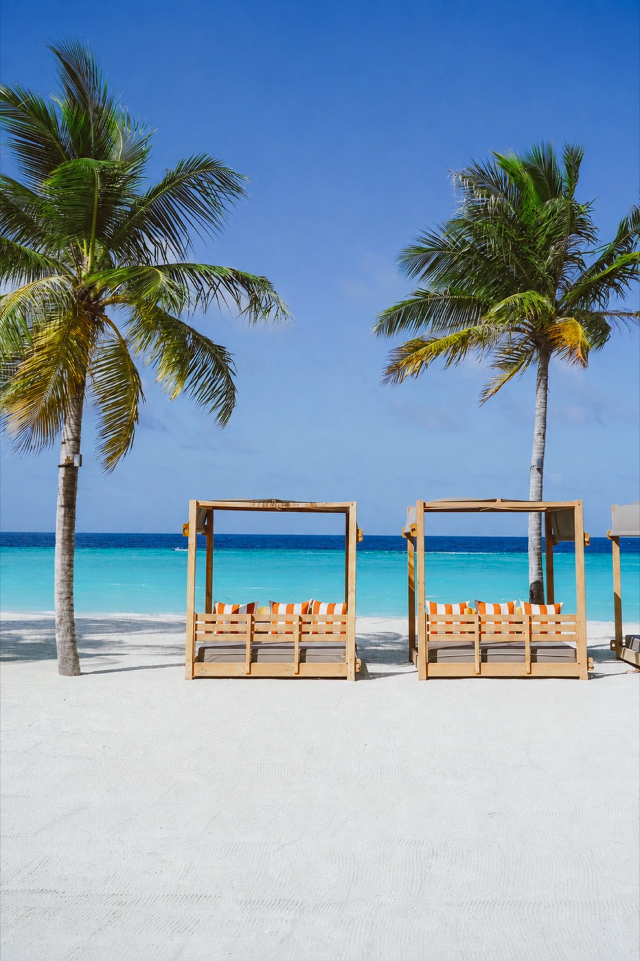 Beach cabana daybeds on white sand at SunRay Maldives, framed by palm trees and turquoise sea