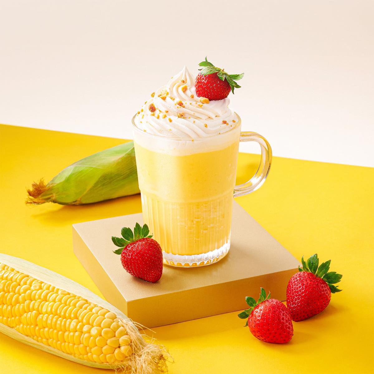K Fry Velvet Sweet Corn Shake in a tall glass, topped with whipped cream and a strawberry, beside fresh corn