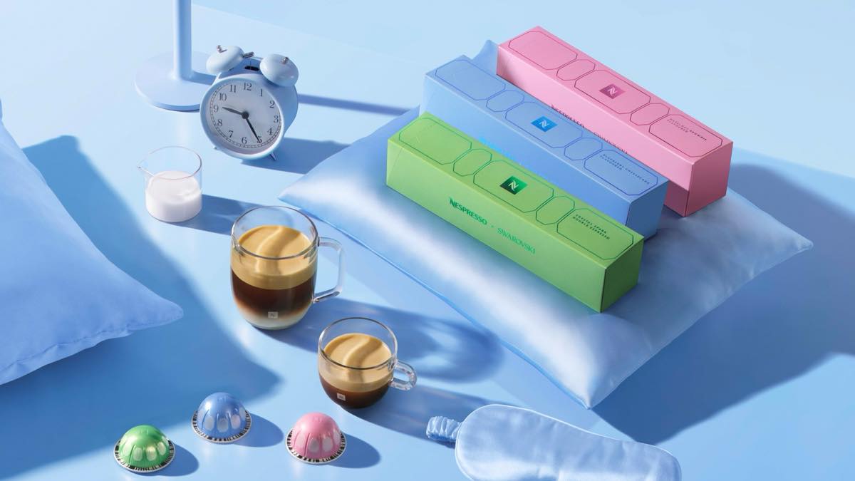 Three Nespresso x Swarovski festive Vertuo coffee boxes in green, blue and pink with matching capsules and two cups of coffee on a soft blue set